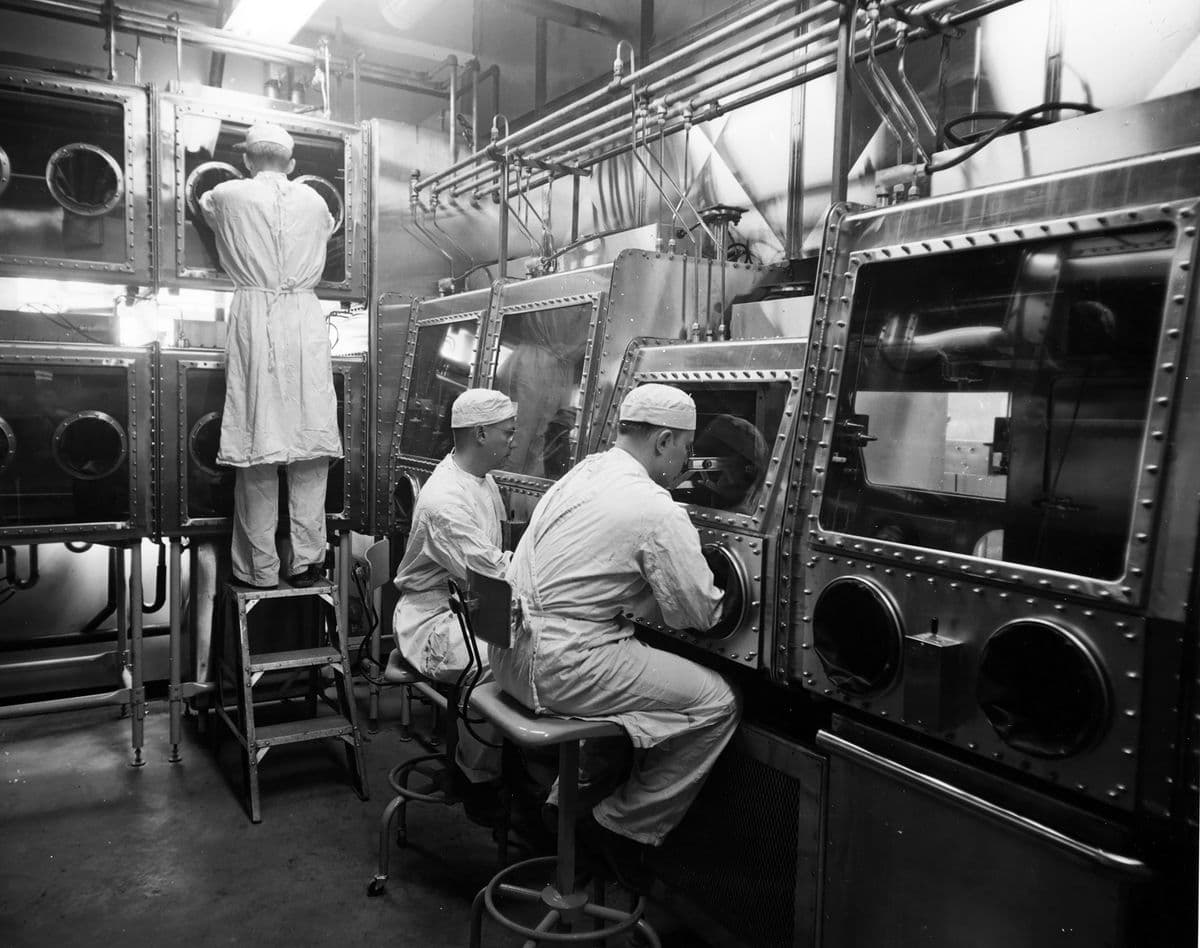 이 이미지는 보호복을 입고 장착된 장갑이 있는 밀폐된 칸막이를 통해 물질을 다루고 있는 세 명의 실험실 작업자들을 보여주고 있습니다.